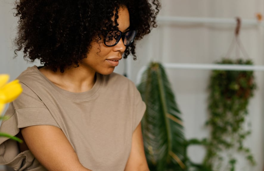 A female professional working in an office with white walls and indoor plants next to a white coat rack donning one empty hanger on it behind where she is sitting at a table of which is not visible, working. The lady with dark frizzy hair and a green t-shirt and thick trendy black framed reading glasses is sitting in a way with her arms slightly extended out in front of her and looking straight and slightly down from her sight line is appearing as if she is working from a computer and typing.