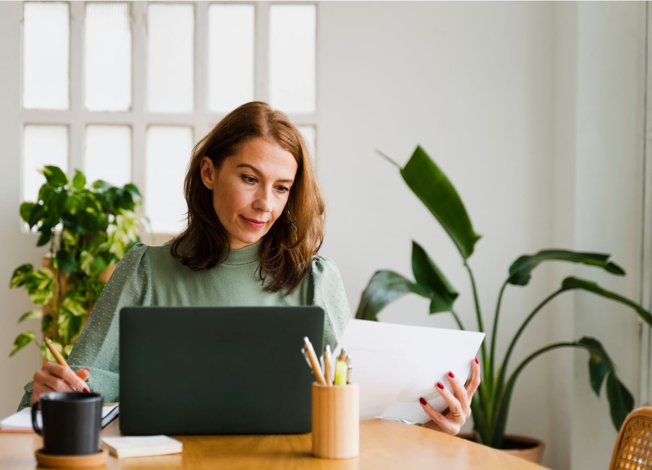 A female professional working in a bright office with white walls and indoor plants behind where she is sitting at a light wood stained table that is serving as her desk. On the desk is a pen cup, two coasters with one of them having a black coffee mug atop of it, a notebook of which she is writing in while hold another piece of paper above the surface of the table in the other hand. The lady with strawberry red hair and a grey sweater top is taking notes from what is on the computer screen as you can clearly see her looking at the laptop while taking notes.
