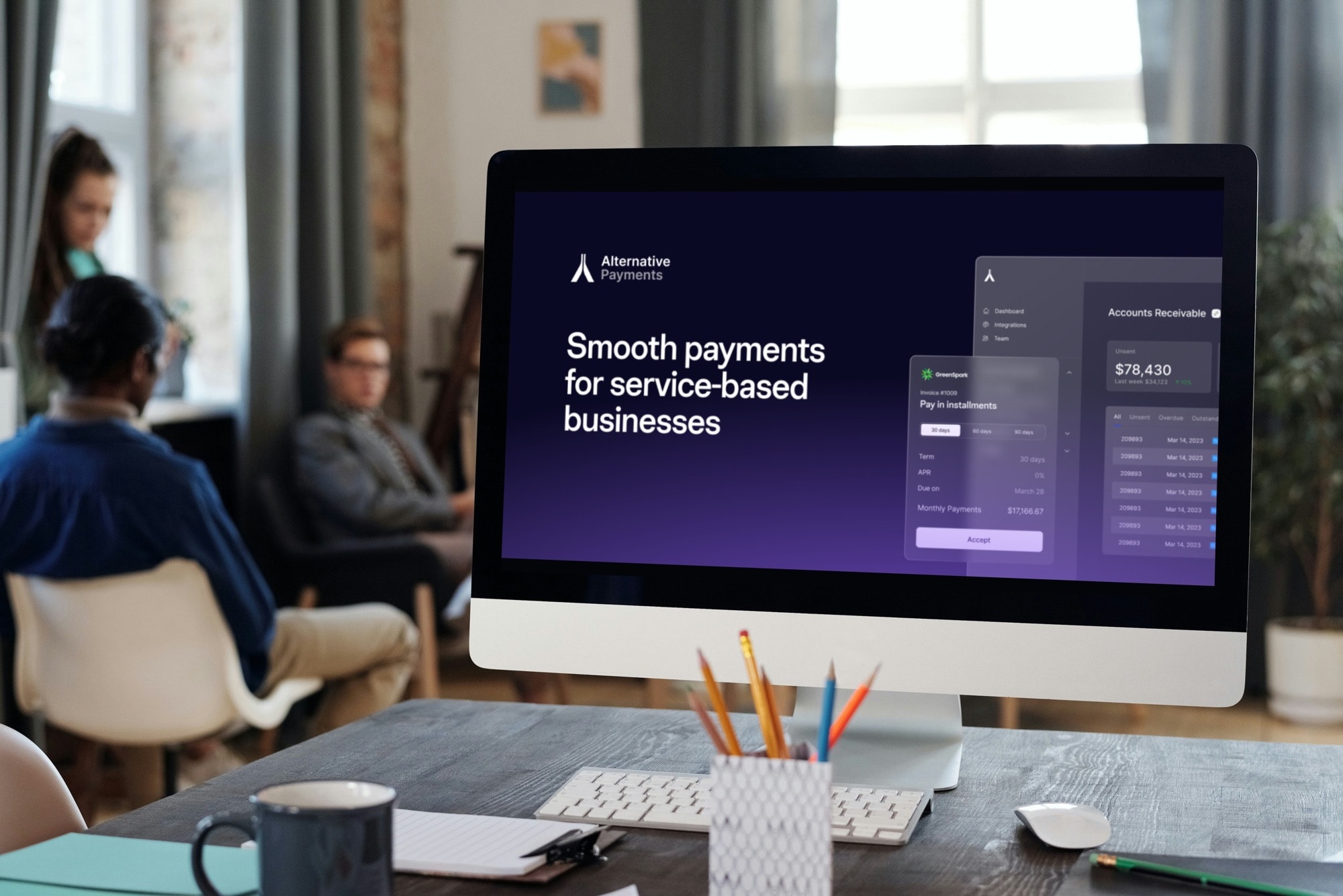 An image of a office setting where there are people sitting in chairs on the outskirt of the photo and in the background. The foreground of the image is a desk with a notepad, pen cup holder, keyboard, mouse, and desktop apple computer. On the computer screen is the homepage for the Alternative Payments website where you can understand how best to choosing the right Payment Service Provider. The website in the image has the wording "Smooth payments for service-based businesses" set on a dark blue, navy, and purple color scheme. Above the computer is a window with light beaming through from the sun outside.
